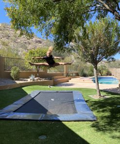 A man joyfully jumps on a trampoline in a sunny backyard, surrounded by green grass and trees.