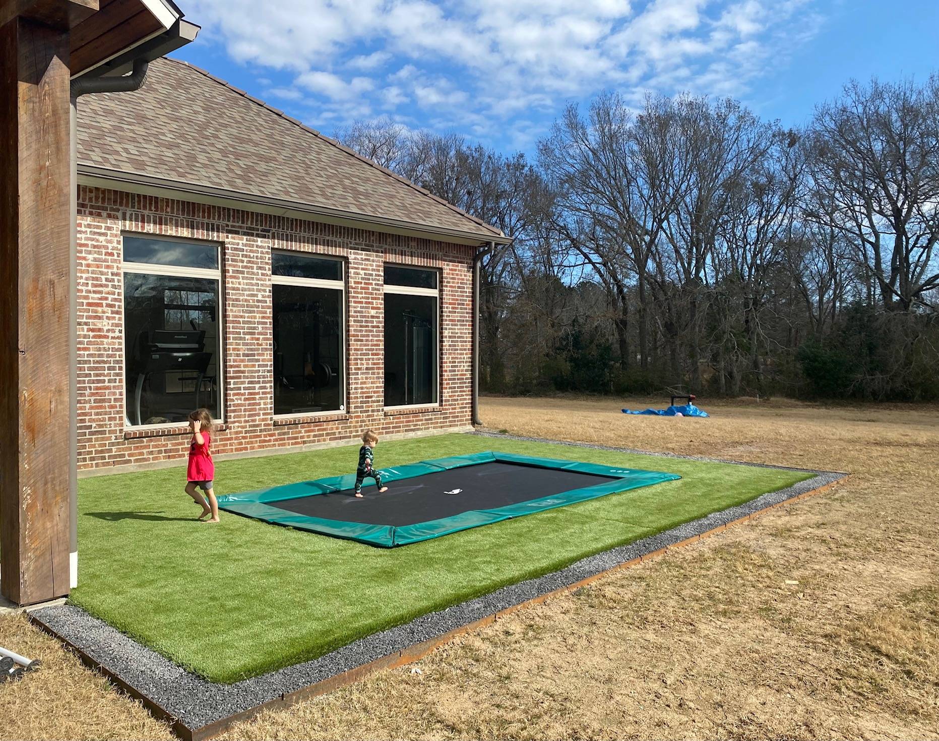 17ft x 10ft Akrobat Primus In-Ground Trampoline - Green (Green Pad with Black Jump Mat) *With Free Enclosure and Cover* 2 A young child happily jumping on a trampoline in a sunny backyard filled with green grass.