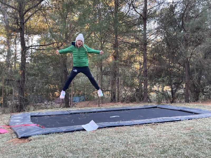 Is Jumping on a Trampoline Better Than Running? 2 A person joyfully jumping on a trampoline in a sunny yard, surrounded by grass and trees.