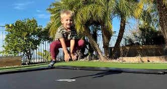 A young boy joyfully jumps on a trampoline in a sunny backyard, surrounded by green grass and trees.